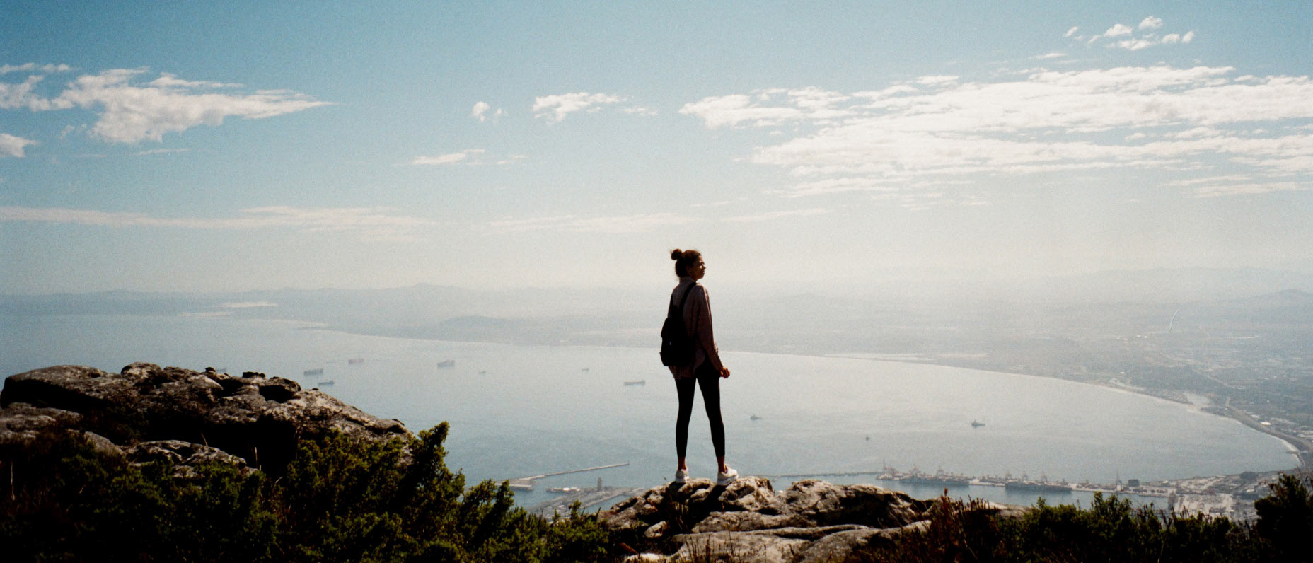Person standing on a hill with view of water and horizon in the distance under cloud cover on the hero banner
